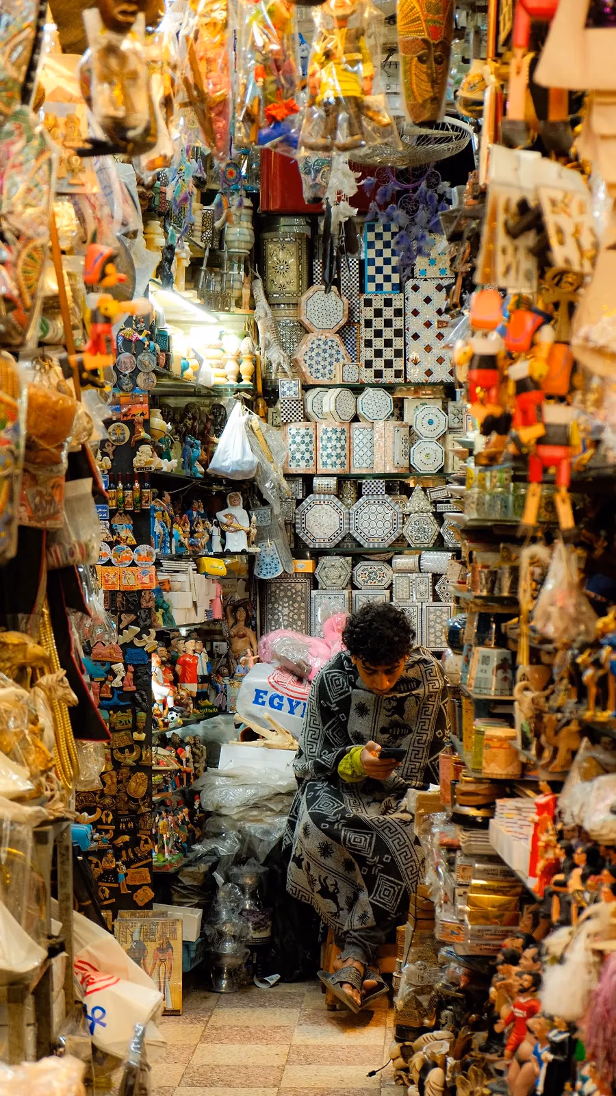 Een kraam in een bazaar met stapels borden, lampen en souvenirs en een verkoper tussen de waren