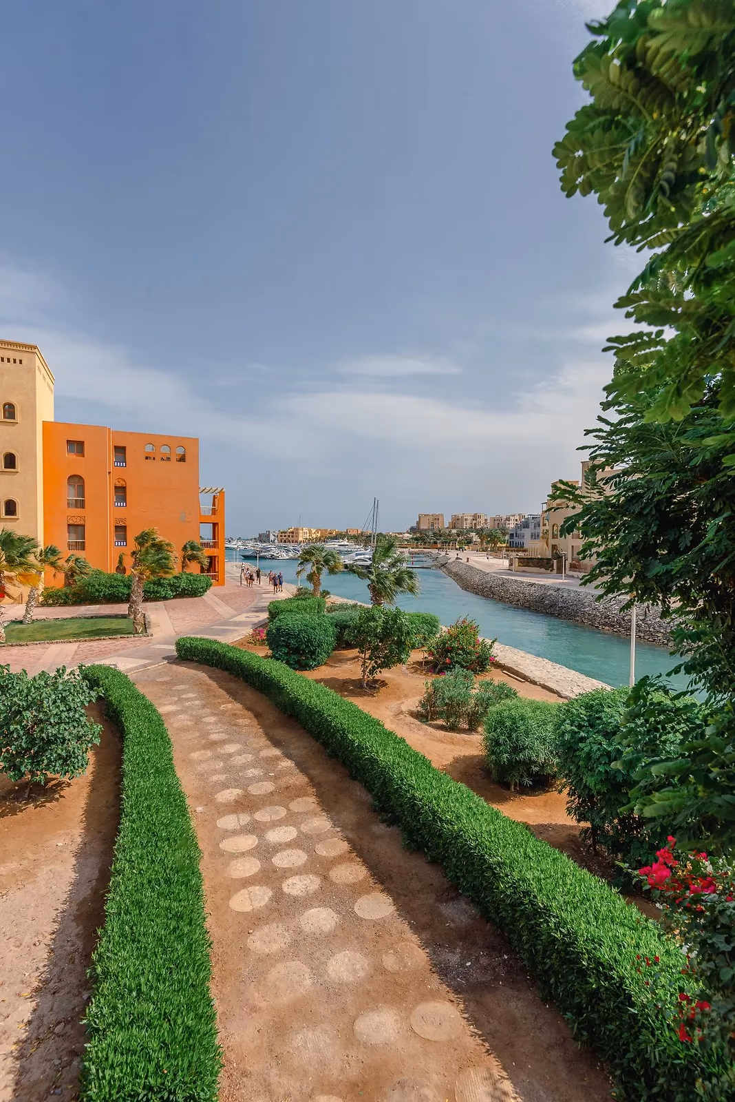 Pad tussen lage hagen langs een kanaal met een oranje gebouw en palmen in El Gouna