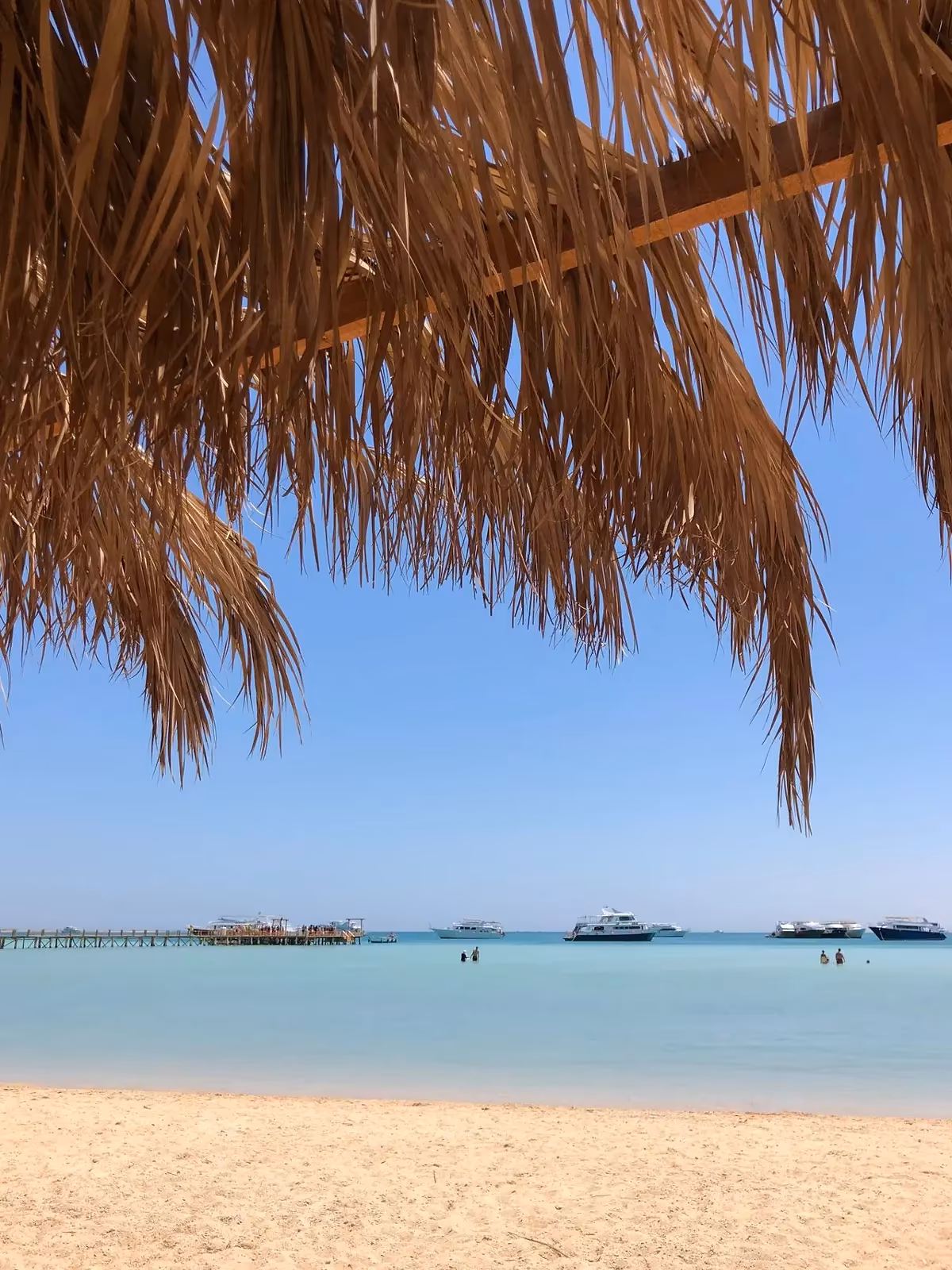 Zandstrand onder het rieten dak van een parasol met ondiep turquoise water en een steiger in de verte