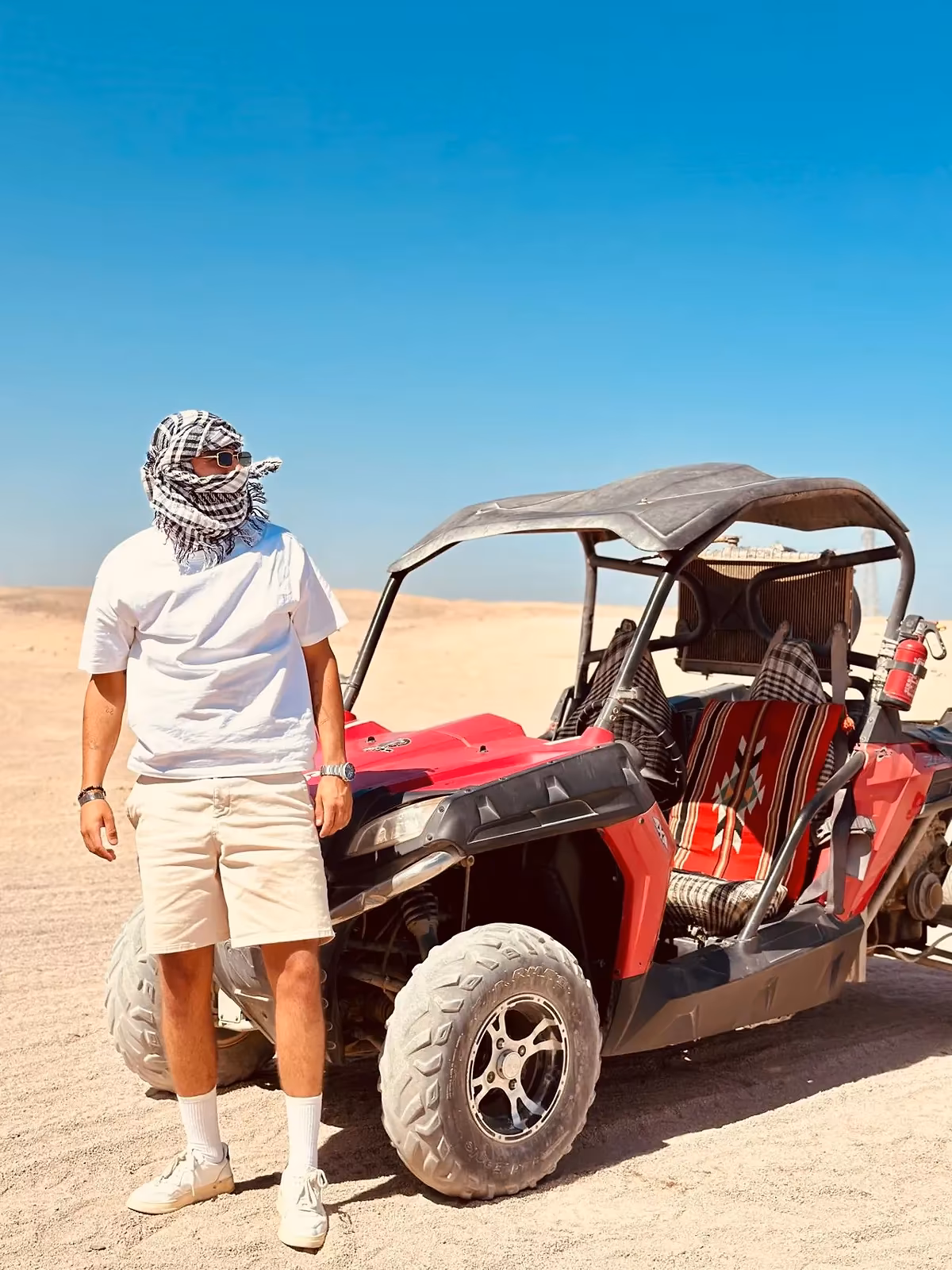 Een man met een kleed om het hoofd naast een rode buggy in het zand van de woestijn