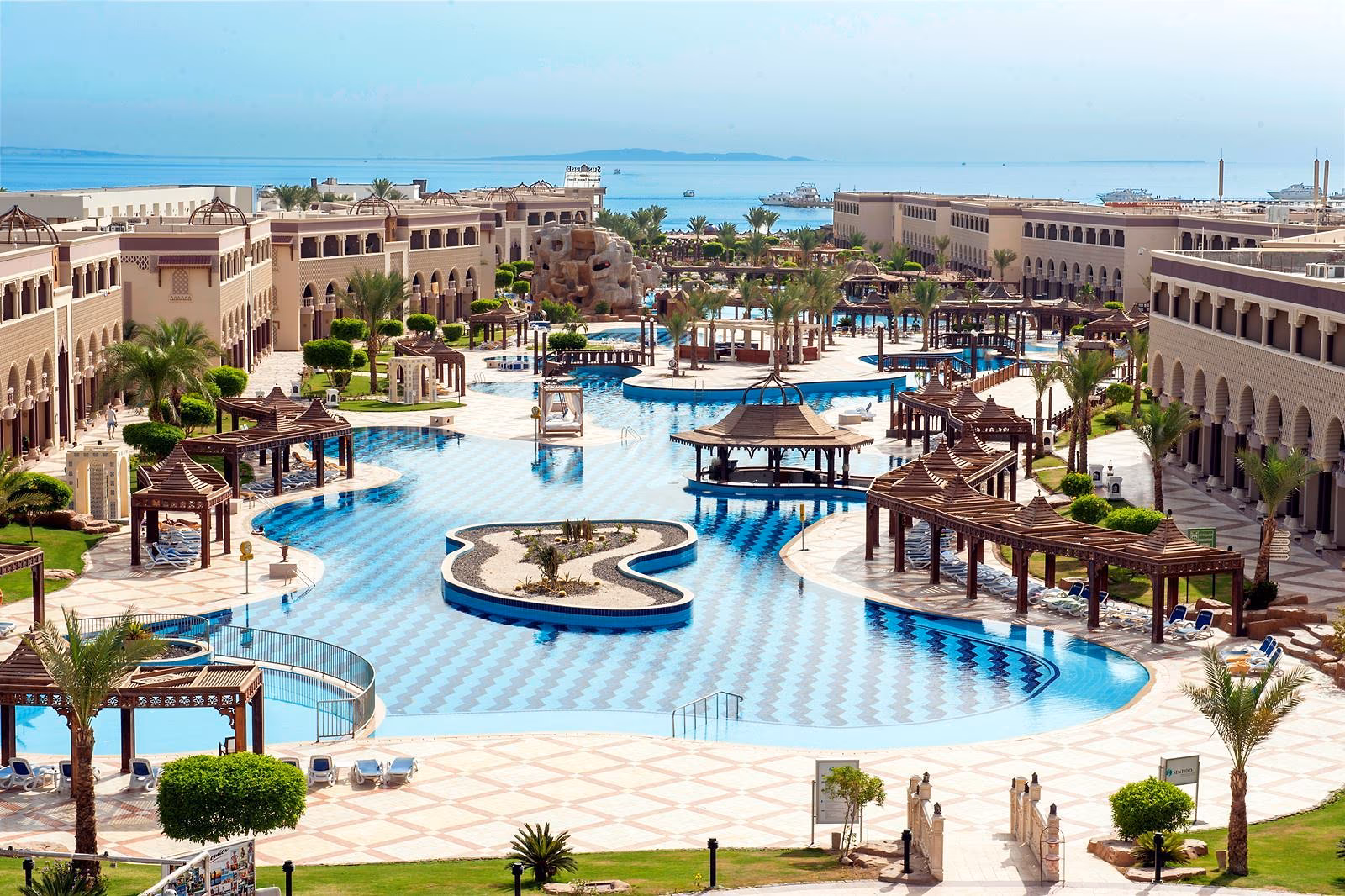 Sentido Mamlouk Palace van bovenaf: zwembaden met een eilandje en houten paviljoens tussen gebouwen met bogen, de zee erachter
