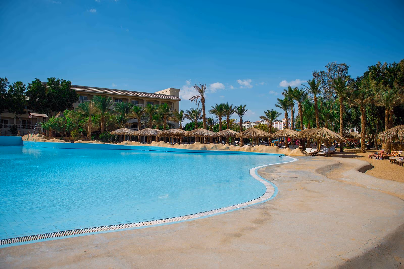 Zwembad met een zandkleurige rand, rieten parasols en palmen bij Sindbad Club